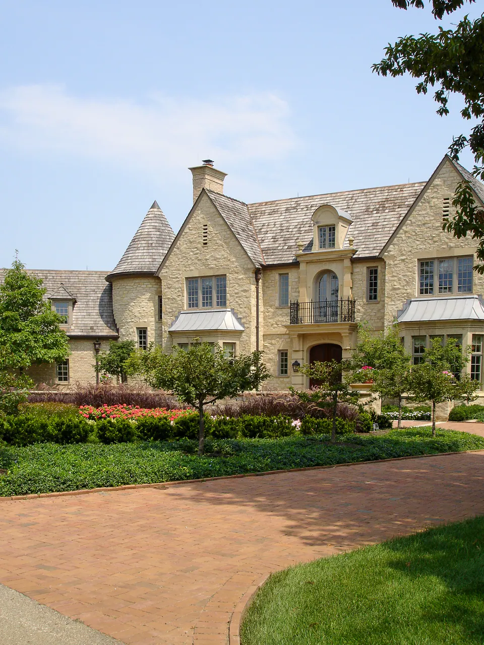 Estate home with brick drive, lawn, hedges, foundation planting, and stone facade partly framed by trees.